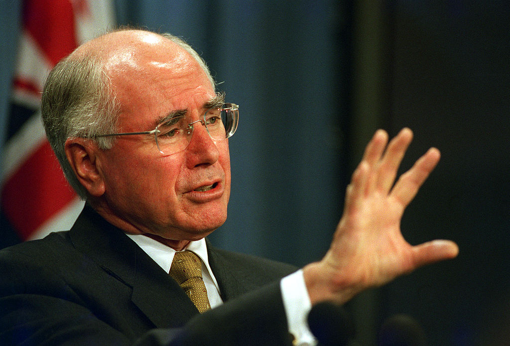 John Howard stands with his hand raised with the Australian flag in the background. 