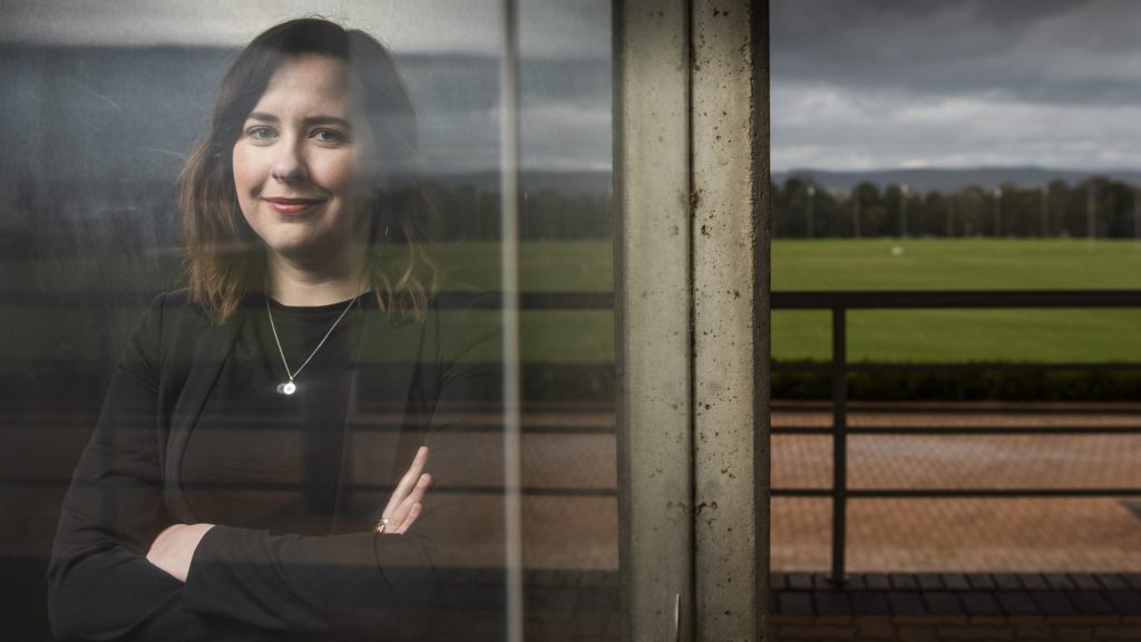 Dr Megan Evans&#039; reflection in a window, with a grassy field behind her. She is smiling and has her arms confidently crossed.