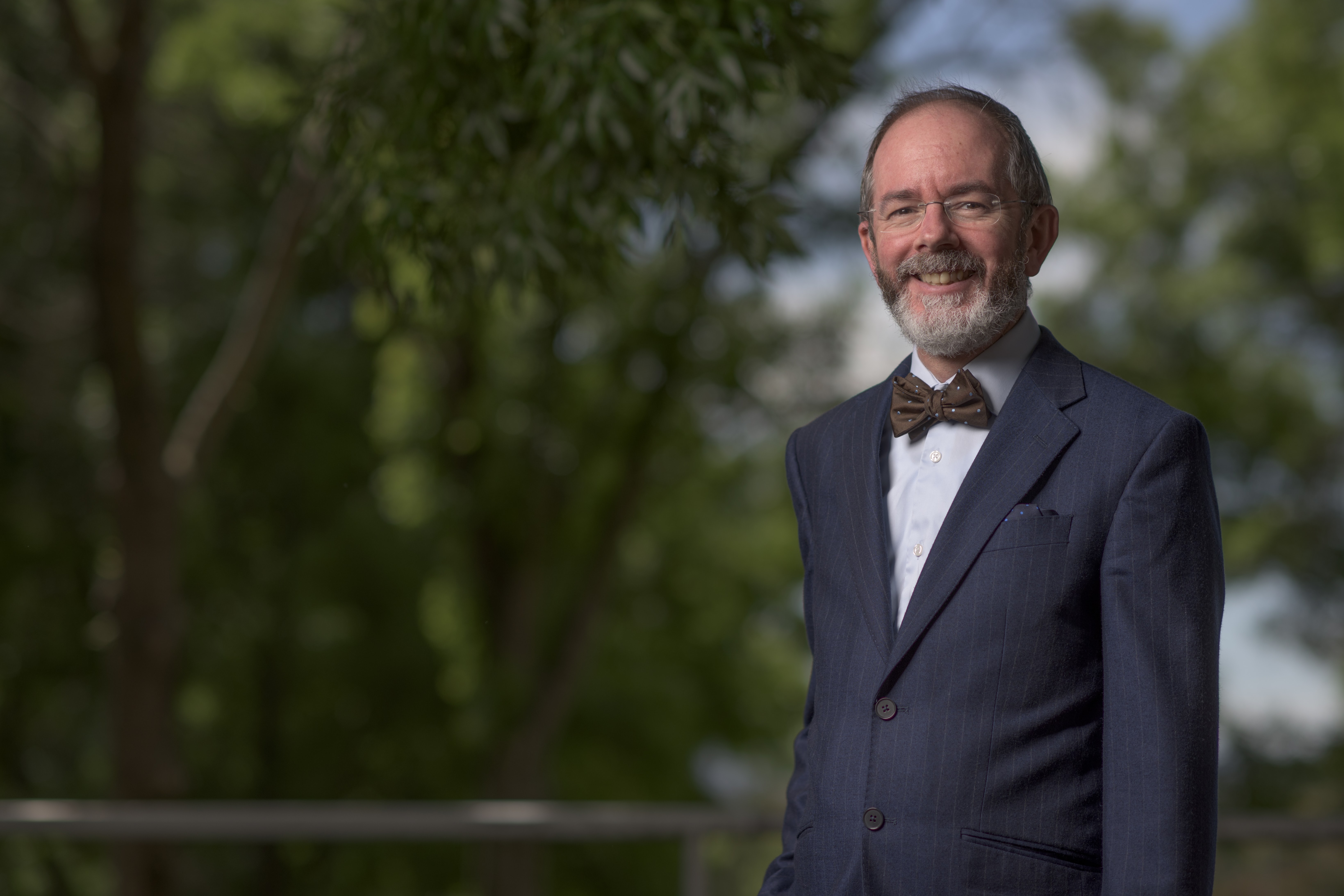 A/Prof. Douglas Guilfoyle is standing outside in front of greenery, wearing a suit and bow tie. He is smiling.