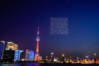A QR code is formed by illuminated drones in the sky in Shanghai, China. The sky is dark, and buildings are illuminated with coloured lights in the background. (Credit: Bilibili)